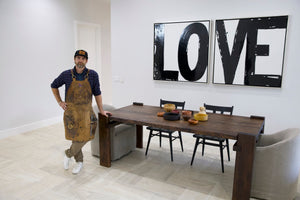 Man in apron standing next to a wooden dining table with 'LOVE' artwork on wall