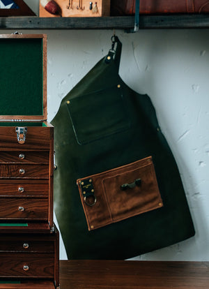 Green apron with a brown pocket hanging on a white wall next to a wooden dresser.